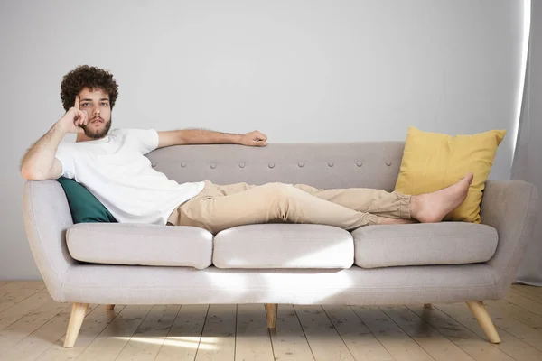 Young man with stubble and voluminous hair lying comfortably on gray sofa in living room.