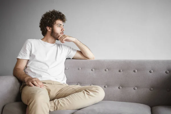 Serious young bearded guy having pensive facial expression, touching stubble, deep in thoughts, pondering. Man sitting on sofa.