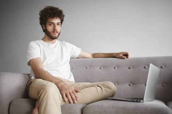 Stylish young man wing thick beard and wavy hair sitting on couch in front of open portable computer, using wireless internet connection.
