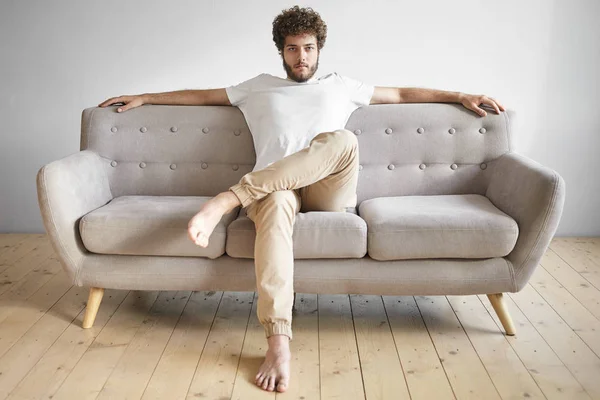 Young man with thick beard having rest at home, sitting casually on luxurious sofa, feeling relaxed.