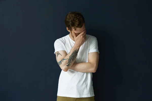 Shy embarrassed young man with beard and tattoos making faceplam gesture.