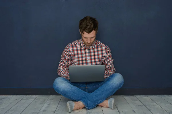 Attractive casually dressed young man with beard sitting on floor with legs crossed, surfing web stores while shopping online using laptop