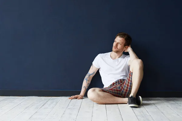 Unshaven man with tattoos wearing black sneakers, t-shirt and shorts having rest indoors.