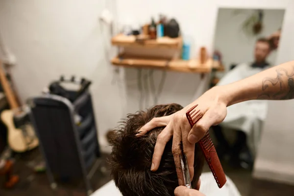 Hairdresser holding comb and scissors cutting hair of male customer who is sitting on chair in front of mirror at barbershop.