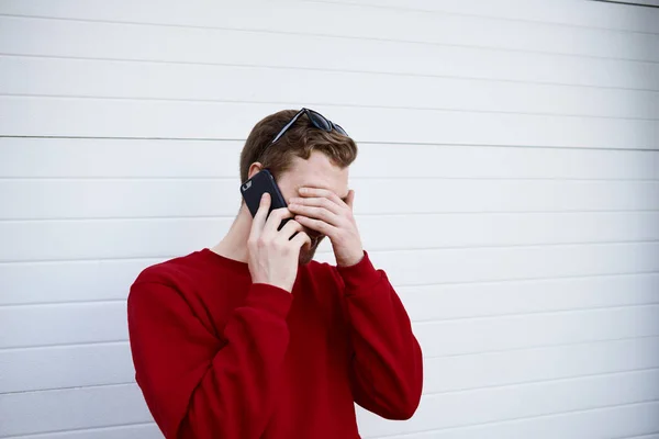 Bearded young man in sweater having phone conversation, making facepalm gesture while laughing at funny story or joke.