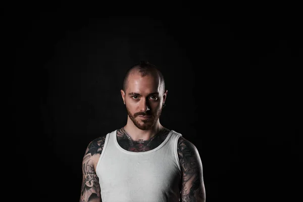 Brutal and brave man in white t-shirt looking at camera. Tattooed strong body person on black background. Tattoo model with short hair style.