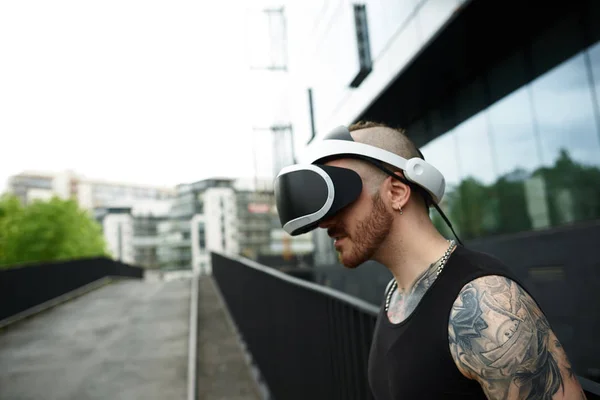 Virtual reality concept. Man wearing VR googles in city. Gaming beard tattooed man near modern business building.
