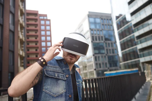 Virtual reality concept. Man wearing VR googles in city. Gaming beard tattooed man near modern business building.