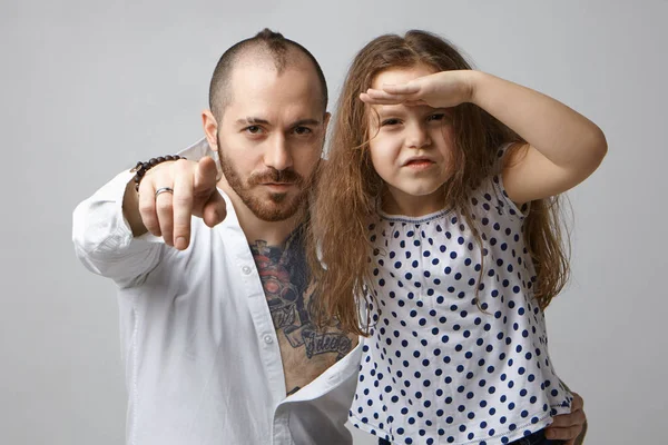 Fashionable young father with beard and tattoo on chest pointing finger at camera, showing something to daughter holding palm at forehead, looking into distance