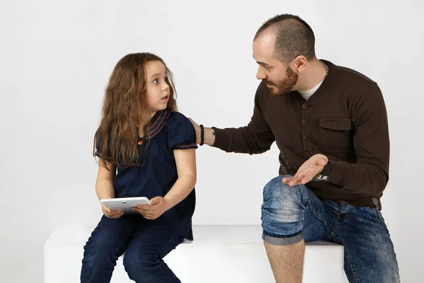 Frustrated female child feeling upset while being told by young father go out and play in real life instead of spending too much time online and surfing internet on electronic tablet.