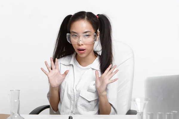 Fascinated bug eyed young asian woman scientist in transparent goggles and white medical coat gesturign actively with open palms, feeling excited about breakthrough scientific research results