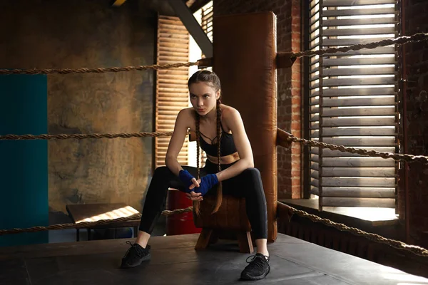 Confident young female fighter wearing handwraps and sports clothes having break during training, sitting in the corner of boxing ring