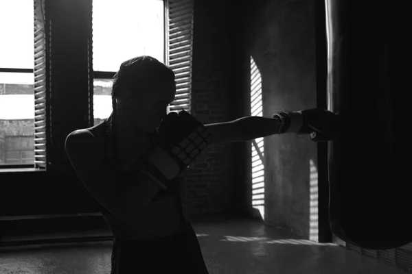 Silhouette of determined professional young female boxer with muscular arms wearing sports outfit and boxing gloves while training in gym, reaching out one hand, punching air in front of her
