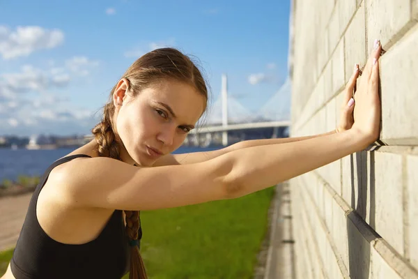 Serious self determined young sportswoman with strong arms posing by brick wall with blue sky and river in background, looking at camera. People, health, activity and sports