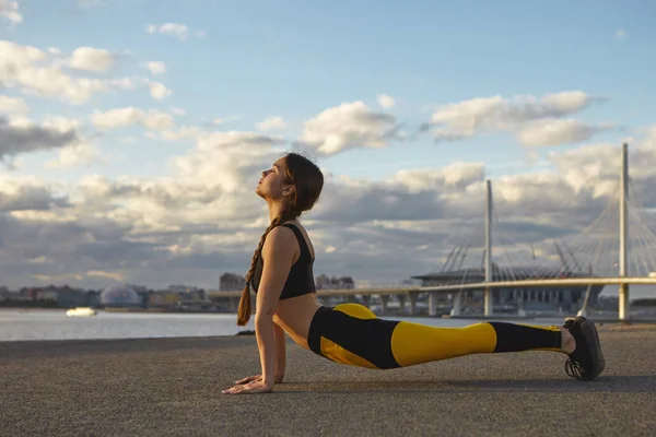 Young sportswoman with strong arms exercising against river background, doing yoga, practicing upward facing pose or Urdhva Mukha Svanasana