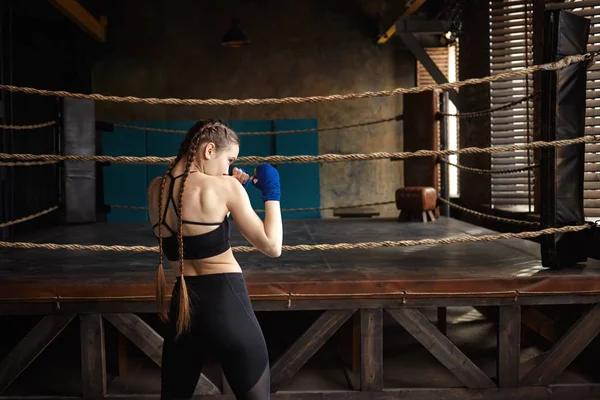 Rear view of professional female boxer with two braids standing in empty gym with boxing ring in background, training alone, working on punching techniques. People, sports and active healthy lifestyle