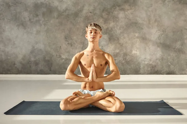 Handsome young guy with blonde hair and tattoo on naked torso sitting on yoga mat in lotus pose, doing Sukhasana, closing eyes and pressing hands together in namaste. Meditation and concentration