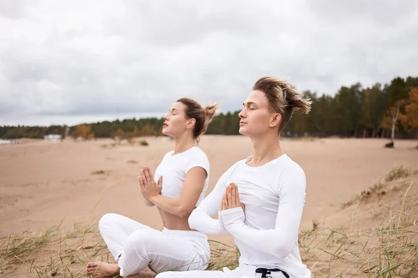 Two atheltic European people man and woman in white sportswear closing eyes and holding hands in namste gesture, sitting in padmasana while meditating outdoors during yoga retreat by ocean