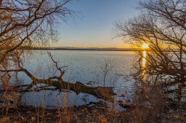 Very peaceful scene of a calm Barr Lake, Barr Lake State Park, in Brighton, Colorado. The image was taken from the shore, and framed with bare trees. There is a log in the lake, just feet from the shore. The sun is setting behind the mountains in the