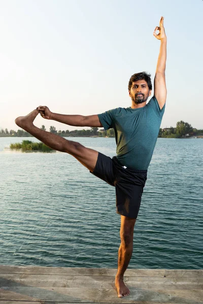 Indian man practicing yoga pose near water standing on wooden path with natural background