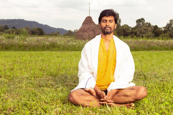 Indian yoga master practicing meditation in lotus postion joined hands with white blanket on expressing calm and relaxation in natural grass background