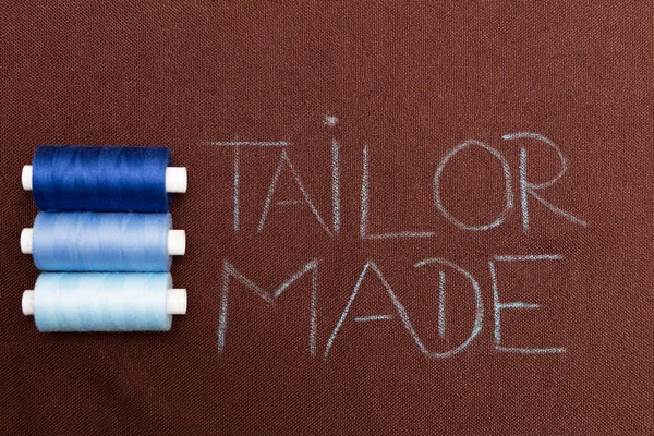 Blue thread bobbins near tailor made text written with chalk on brown material