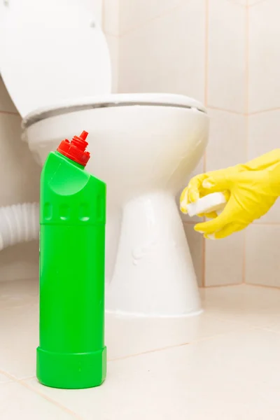 Green disinfectant bottle and white sponge held with yellow sanitary gloves in front of wc as chores concept