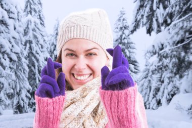 Kadın umutlu ifade parmak tutan kış kıyafetleri ile iyi şanslar jest mor eldiven olarak kar ve ağaçlar arka plan üzerinde geçti