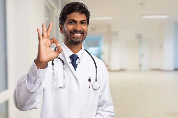 インドの医師男笑顔 クリニック廊下背景で大丈夫のジェスチャーとして指を押し — ストック写真