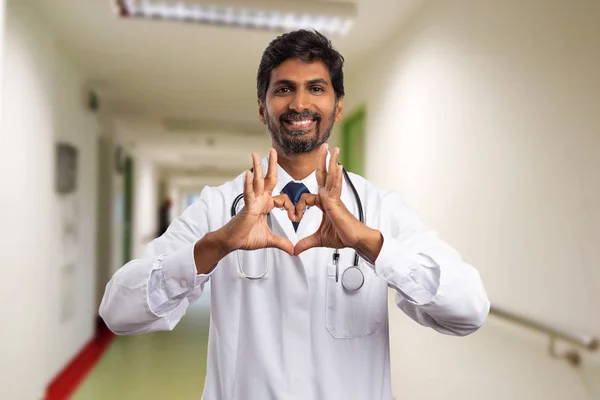 Indian physician or medic smiling while making heart symbol with fingers as I love my job concept in hospital hallway
