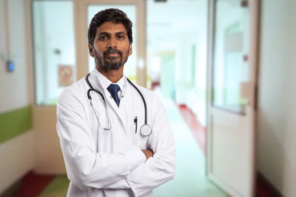 真剣な表情と組んだ腕を背景としてクリニック廊下に開いているドアを持つ白のコートを着ている男性のインド医師の肖像画 — ストック写真