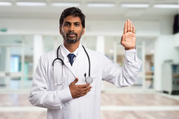 Indian male medic or doctor swearing with hand on heart and palm raised as solemn hippocratic oath concept in hospital hallway background