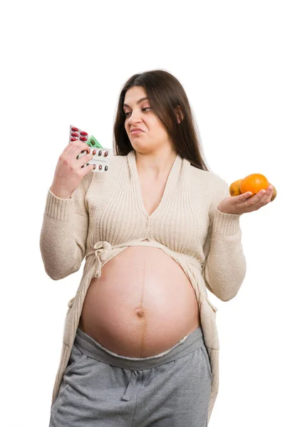 年轻的怀孕妇女看上去对药物感到厌恶 并持有白色背景的水果 认为这是不健康的选择概念 — 图库照片