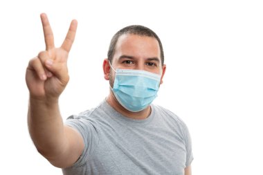Tek kullanımlık koruyucu cerrahi maske takan beyaz adam stüdyo arka planında izole edilmiş koronavirüs gribi salgını sırasında iki parmağıyla barış ya da zafer işareti yapıyor.
