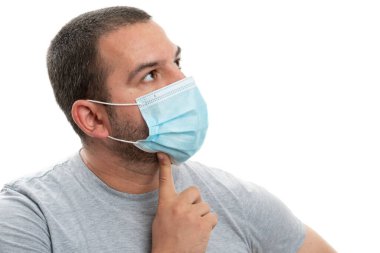 Yetişkin adam boş telif alanına bakıyor. Beyaz üzerine izole edilmiş 19 salgın önleme kavramı olarak cerrahi maske takan işaret parmağıyla çeneye dokunmak için.