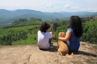 Genç bir kadın kıvırcık, kıvırcık bir çocuk ve bir köpek dağlarda yüksekliğinde bir kaya üstünde oturuyor 
