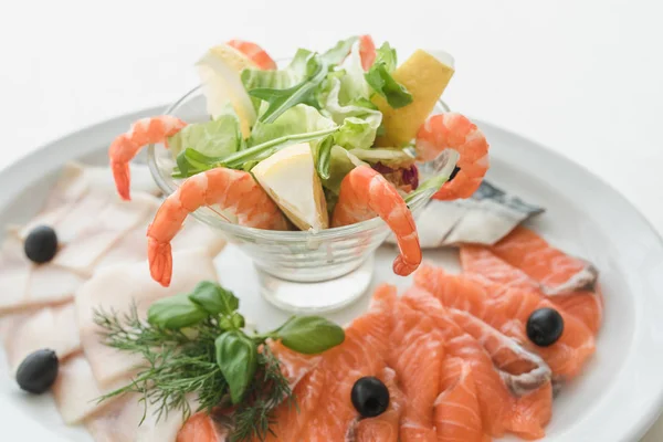 Fish dish with red and white fish, tiger shrimps, served with lemon and lettuce leaves. The dish is decorated with basil and dill, as well as olives. A plate on a table with a white tablecloth.