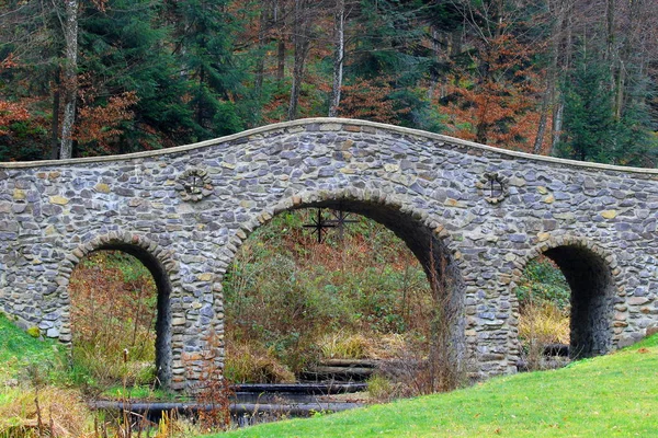 Ancient stone arch bridge crossing a small stream among the green trees of a park in the mountains of the Carpathians, a wild mountain landscape.