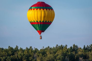 Sıcak hava balonu mavi gökyüzünün arka planında uçuyor. Geniş yeşil yaprak döken bir ormanın üzerinde uçma anı. Renkli balon suluboya resim.