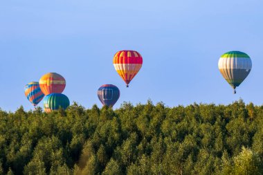 Bir düzine parlak renkli balon mavi gökyüzünün arka planına doğru uçuyor. Geniş yeşil yaprak döken bir ormanın üzerinde uçma anı. Çok renkli balonlar farklı mesafelerde yüzerler..