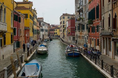 Venedik, İtalya - 16 Ekim 2024: Renkli binalar Venedik, İtalya 'da insanların su boyunca yürüdüğü dar bir kanal çiziyor. Küçük bir taş köprü zarif bir şekilde en uçta kıvrılır..