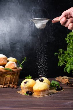 Siyah zemin üzerinde pudra şekerli nane ve kuşüzümü yapraklı kapkekler, içinde kekler ve kuşüzümü olan ahşap bir sepet. Keklerin üzerine pudra şekeri konması.