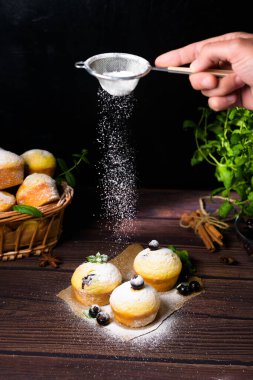 Siyah zemin üzerinde pudra şekerli nane ve kuşüzümü yapraklı kapkekler, içinde kekler ve kuşüzümü olan ahşap bir sepet. Keklerin üzerine pudra şekeri konması.