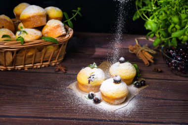 Siyah zemin üzerinde pudra şekerli nane ve kuşüzümü yapraklı kapkekler, içinde kekler ve kuşüzümü olan ahşap bir sepet. Keklerin üzerine pudra şekeri konması.