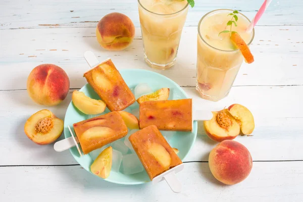 peach ice cream on a stick in a blue plate and ice cubes, peach refreshing juice in a glass with ice and pieces of fruit and mint in the summer. top view on a light background.