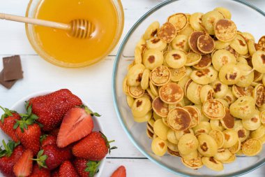 Mini krep gevreği kasesi taze çilekli muz çikolatası, hindistan cevizi talaşı ve küçük tabakta bal. Yukarıdan manzara. Kendini soyutlama eğilimi.