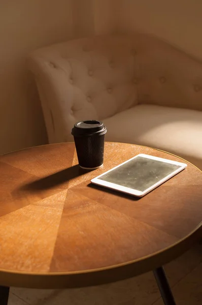 Eine Einwegtasse Kaffee und ein digitales Tablet auf einem Holztisch im Innenraum mit Sonnenstrahl — Stockfoto