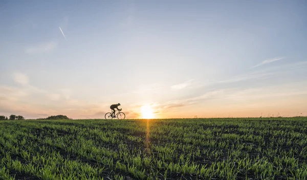 Landbouwgronden en een man fietstocht een in de zonsondergang. Fietsen bij zonsondergang. Gezonde levensstijl Concept. Mannelijke rit fiets in de zonsondergang. Biker fietsen op zonsondergang hemel op een veld. Sport. — Stockfoto