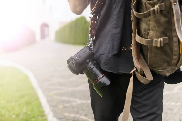 Back of professional wedding photographer in black shirt and with a two cameras an a shoulder straps and brown bag against empty road in green garden. Wedding photography.