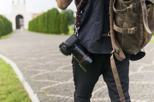 Costas de fotógrafo de casamento profissional em camisa preta e com duas câmeras um um ombro tiras e bolsa marrom contra estrada vazia em jardim verde. Fotografia casamento . — Fotografia de Stock